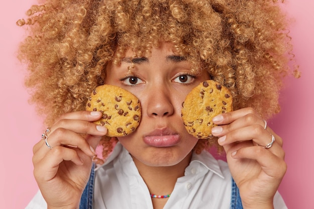 Free photo horizontal shot of gloomy sad woman looks unhappily at camera holds two delicious cookies over face tries to keep healthy diet feels temptation to eat yummy sweet food isolated over pink background