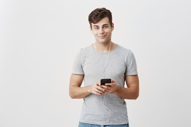 Horizontal shot of handsome caucasian guy dressed casually, holds modern smart phone, listens melodies in earphones, enjoys free internet connection, has happy expression, poses in studio