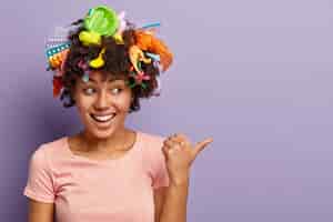 Free photo horizontal shot of happy dark skinned woman with litter in hair, points thumb aside, demonstrates copy space, laughs positively, being active volunteer, wears casual t shirt. garbage and recycling