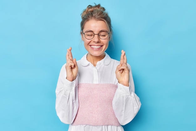 Free Photo horizontal shot of happy elegant woman in formal shirt keeps eyes closed crosses finger believes in good luck isolated over blue background. big hope for dreams come true. body language concept