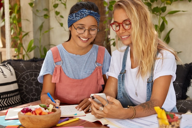 Free photo horizontal shot of happy females discuss funny blog in internet, use mobile phone