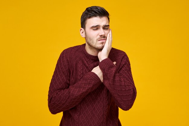 Horizontal shot of happy frustrated young male in knitted jumper having problems with tooth cavity needs to see dentist, keeping hand on his cheek and grimacing, canât stand terrible toothache