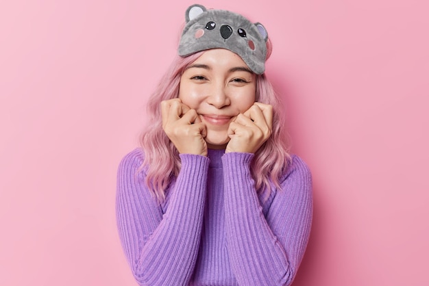 Horizontal shot of happy rosy haired Asian woman keeps hands under chin has gentle expression looks glad at camera wears sleepmask casual purple jumper isolated on pink background going to have sleep