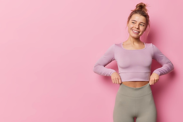 Free photo horizontal shot of happy sporty woman dressed in tracksuit has fit healthy body looks away has cheerful expression poses against pink background with copy space for your advertising content.