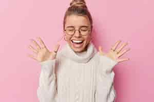 Free photo horizontal shot of happy woman with combed hair raises palms wears round spectacles and white sweater feels amused poses against pink background hears amazing good news screams from happiness