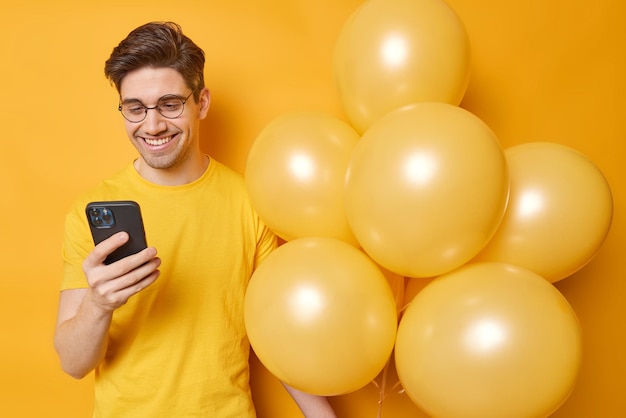 Horizontal shot of happy young man checks notification on smartphone celebrates birthday comes on party dressed in casual t shirt poses with balloons isolated over yellow background Celebration