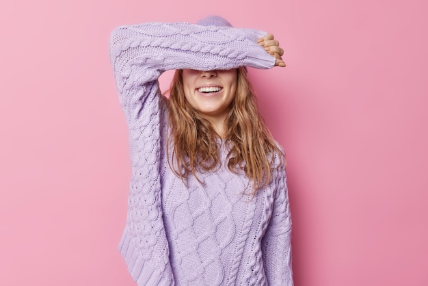 Free Photo horizontal shot of happy young woman covers eyes with arm wears loose knitted sweater tries to hide face stays positive isolated over pink background cheerful female model awaits for surprise