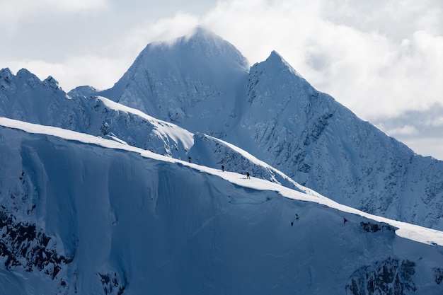 Horizontal shot of high mountains covered with snow under white clouds and a group of people hiking