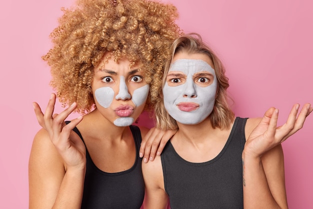 Free photo horizontal shot of indignant women look puzzled raise palms stand closely to each other apply clay masks for rejuvenation and skin treatment dressed in casual black t shirts isolated over pink wall