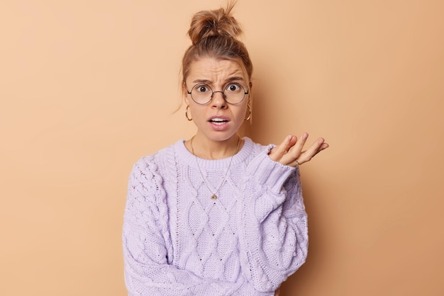 Free photo horizontal shot of indignant young european woman with combed hair looks puzzled shrugs shoulders feels confused wears round spectacles and casual jumper isolated over beige background so what