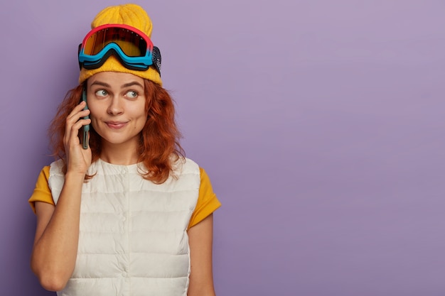 Free Photo horizontal shot of lovely young ginger woman on ski resort, makes telephone call, wears yellow hat and white vest, stands indoor over purple wall