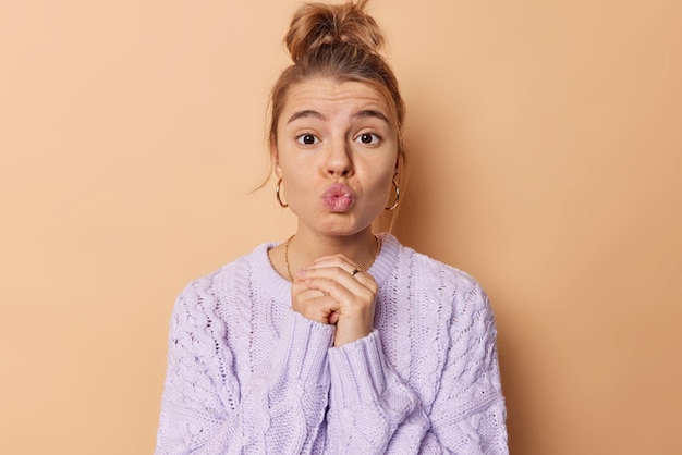 Free Photo horizontal shot of lovely young woman keeps lips folded feels romantic keeps hands together wants to kiss someone looks with tender expression dressed in knitted jumper isolated over beige wall