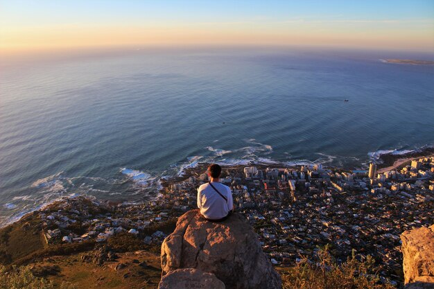 Horizontal shot of a man sitting on the edge of the rock and looking at the coastline city