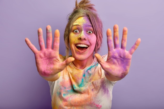 Free Photo horizontal shot of optimistic joyful young girl shows two colorful palms, celebrates holi festival, laughs gladfully, plays with special colored powder. focus on painted hands. splash of color