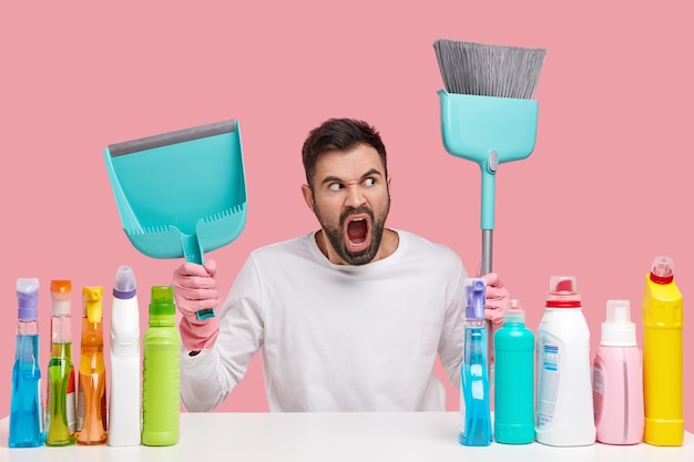 Horizontal shot of outraged man has thick beard, carries broom and scoop, shouts angrily, dressed in white clothes, angry with much work