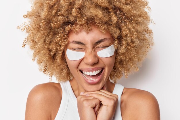 Horizontal shot of overjoyed curly haired young woman keeps hands under chin applies white beauty patches under eyes to remove bags or wrinkles poses indoor. Morning skin care routine concept