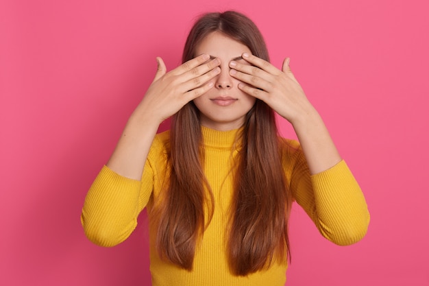 Free photo horizontal shot of peaceful good looking cute young model standing isolated over pink wall in studio, covering eyes with hands, wearing yellow sweatshirt, being alone. surprise concept.