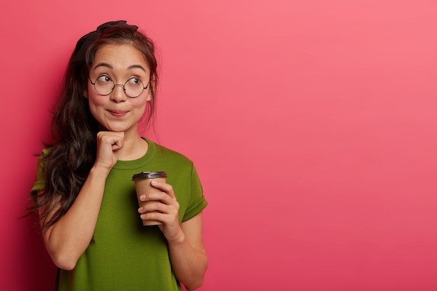 Free Photo horizontal shot of pensive asian schoolgirl dreams about vacation, holds fist under chin, drinks takeaway coffee, enjoys break, stands indoor against rosy wall, wears spectacles and t shirt.