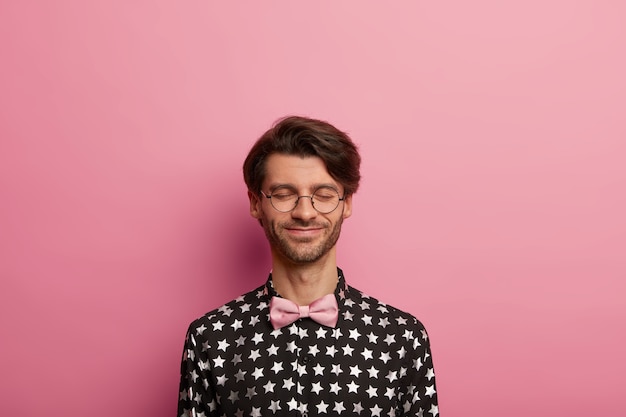 Horizontal shot of pleased hipster guy stands with eyes closed, thinks about something positive, has smile on face, dark bristle