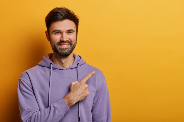 Free photo horizontal shot of positive bearded man indicates at blank space, smiles joyfully, shows nice advertisement, wears violet sweatshirt, isolated over yellow wall. people and promo concept