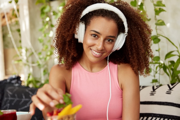 Horizontal shot of positive woman listens new music in modern headphones while waits for friend in cafe, eats fruit dessert, enjoys good rest. African American student recreate after lectures