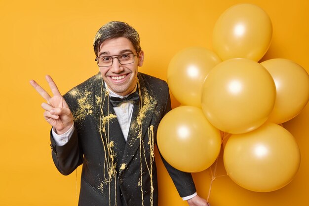 Horizontal shot of positive young man makes peace gesture celebrates birthday has fun on party dressed in elegant formal suit holds bunch of inflated balloons isolated over yellow background