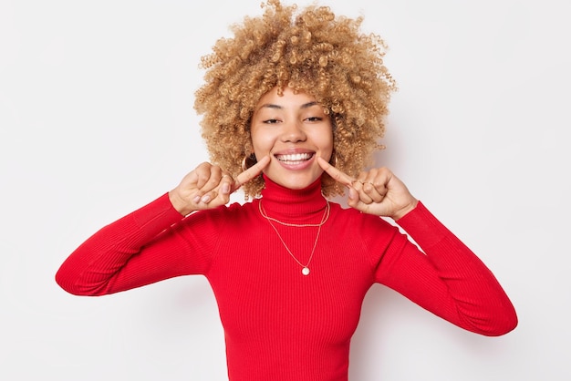 Free photo horizontal shot of positive young pretty woman with curly hair points index fingers at toothy smile shows white even teeth dressed in red turtleneck poses indoor look at my happy perfect smile