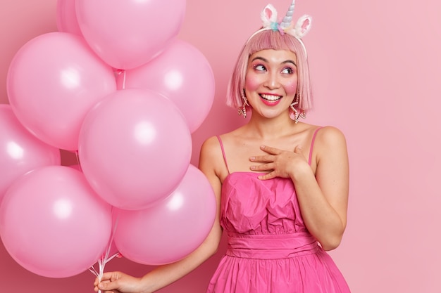 Horizontal shot of pretty cheerful birthday girl accepts congratulations smiles pleasantly holds helium balloons