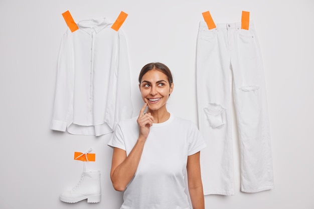 Horizontal shot of pretty young brunette woman dressed in casual t shirt thinks about donation poses indoor against white background with plastered items of clothes has pleasant thoughts in mind