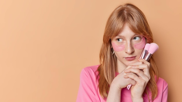 Free photo horizontal shot of redhead girl with freckled skin applies hydrogel pink patches under eyes holds cosmetic brushes focused away isolated over beige background empty space for your promotion