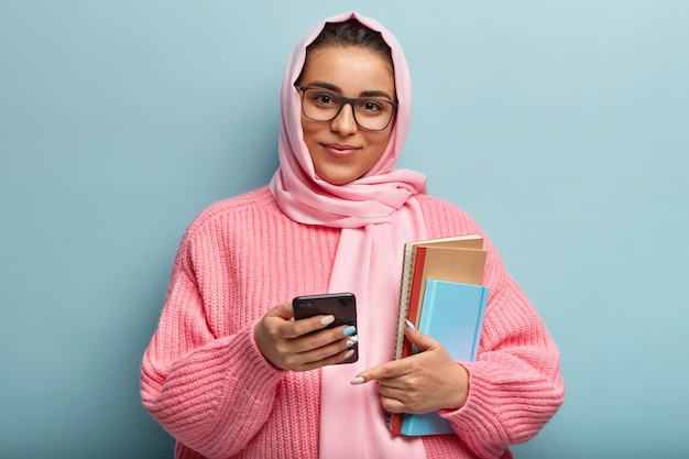 Free photo horizontal shot of satisfied college student uses new cool app on cell phone, carries notepad for writing notes, wears spectacles, silk scarf and knitted sweater, isolated over blue wall.