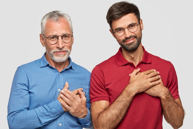 Horizontal shot of satisfied two men of different age, make gratitude gesture, feel thankful to generous people, have pleased expressions, isolated over white wall. Generation, body language