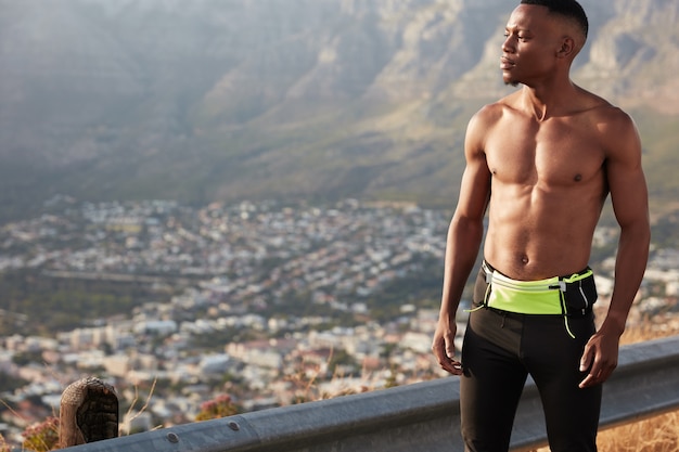 Horizontal shot of self assured sporty man with muscular body, dressed in active wear, has morning workout out of town, focused at distance with free space