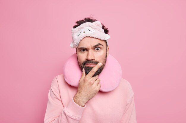 Horizontal shot of serious bearded adult man looks with strict expression wears comfortable travel pillow and sleepmask dressed casually 