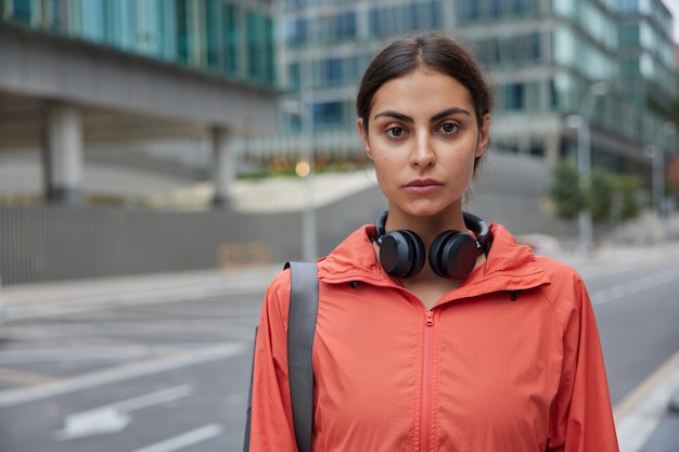 Free photo horizontal shot of serious good looking sportswoman dressed in windbreaker uses wireless headphones returns from workout session after physical activities poses against blurred city