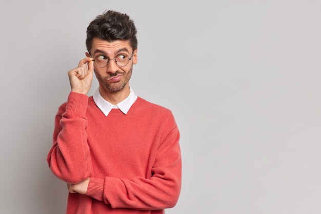 Horizontal shot of serious man looks thoughtfully away keeps hand on rim of spectacles purses lips concentrated on right