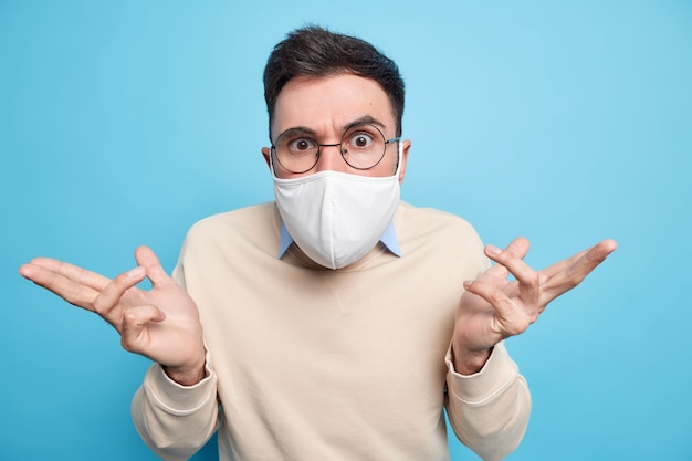 Horizontal shot of serious man wears round spectacles protctive face mask against coronavirus disease spreads hands feels confused cannot make choice 