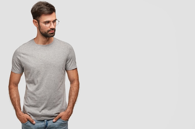 Horizontal shot of serious unshaven male in casual grey t-shirt, keeps hands in pockets, looks aside, thinks about something