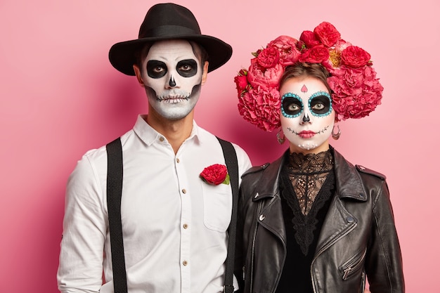 Horizontal shot of serious woman and man dressed in Halloween costumes, wear skeleton makeup, wreath made of peonies