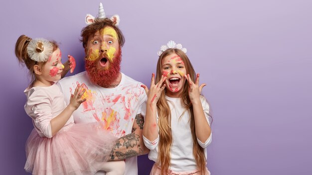 Horizontal shot of shocked dad has yellow face painted with watercolors, two children have fun with father, joyful expressions, isolated over purple wall with free space for promotion.