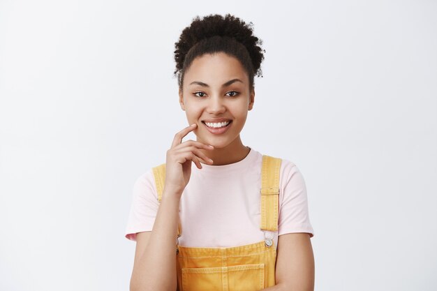 Horizontal shot of smart and creative stylish African American with combed hair in yellow dungarees, smiling broadly and touching cheek while having great suggestion or plan, intrigued over grey wall