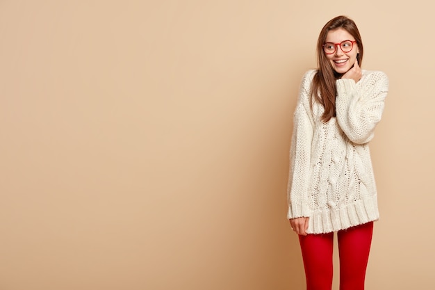 Horizontal shot of smiling glad woman looks on left copy space, expresses sincere feelings, sees something pleasant and funny, wears long knitted sweater and red tights, isolated on beige  wall