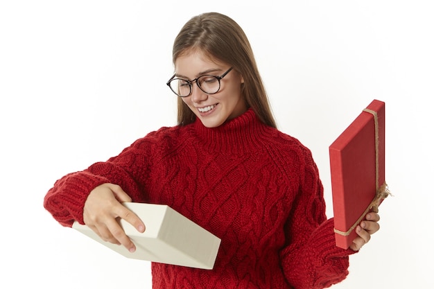 Free photo horizontal shot of stylish joyful young caucasian woman in eyeglasses and knitted sweater having excited happy look while receiving gift for christmas, opening box with present and smiling broadly