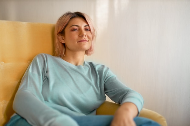 Free photo horizontal shot of stylish pretty girl with bob pink hairstyle and nose ring sitting comfortably in armchair, looking at camera with carefree relaxed smile.