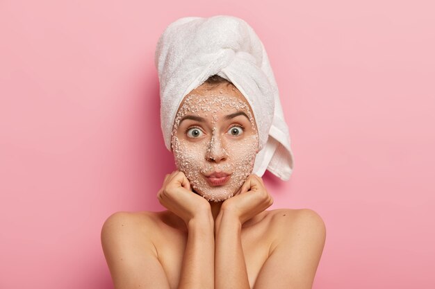 Horizontal shot of surprised Caucasian woman keeps palms under chin, looks with widely eyes, applies scrub mask, avoids problems with skin, stands shirtless against pink wall. Beauty concept