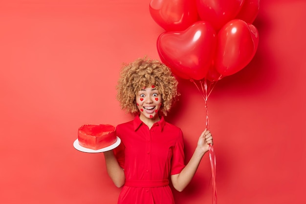 Free photo horizontal shot of surprised positive young woman gets present from boyfriend on valentines day holds heart shaped cake and balloons wears dress enjoys favorite holiday isolated over red studio wall