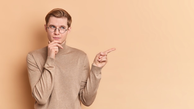 Free photo horizontal shot of thoughtful young man holds chin thinks about suggested offer points away shows copy space for your advertising content wears round spectacles casual jumper