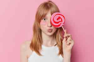 Free photo horizontal shot of young girl with natural ginger haair freckled skin covers caramel candy over eye applies hydrogel patches for reducing puffiness isolated over pink background beauty concept