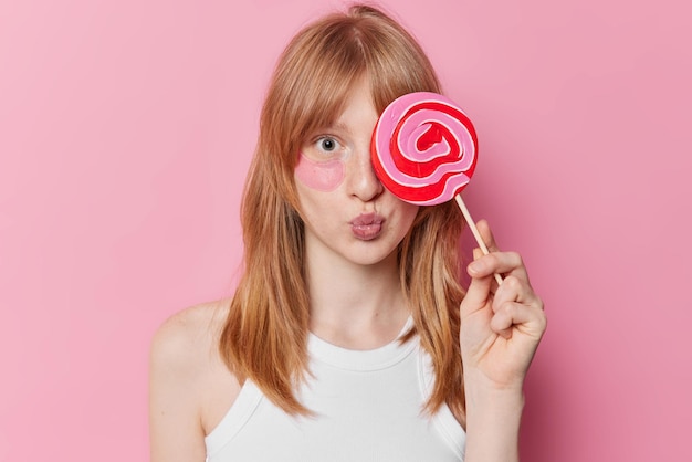 Horizontal shot of young girl with natural ginger haair freckled skin covers caramel candy over eye applies hydrogel patches for reducing puffiness isolated over pink background Beauty concept