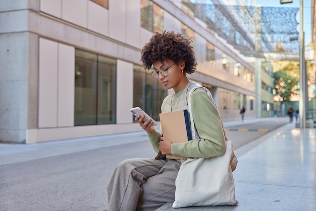 Horizontal shot of young woman sits outdoors looks seriously at smartphone scrolls social networks dressed in casual clothes carries fabric bag rests in city after walking Street lifestyle